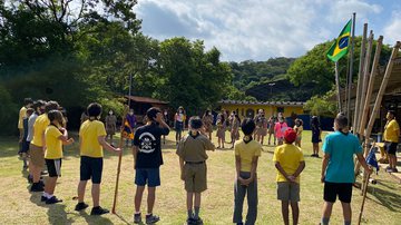 #DescreviPraVocê: foto mostrando a Tropa Escoteira do Grupo Escoteiro Falcão Peregrino em formação de “ferradura” durante cerimônia de Hasteamento da Bandeira Nacional em um conjunto de mastros feitos em bambu. - Fotos: divulgação