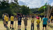 #DescreviPraVocê: foto mostrando a Tropa Escoteira do Grupo Escoteiro Falcão Peregrino em formação de “ferradura” durante cerimônia de Hasteamento da Bandeira Nacional em um conjunto de mastros feitos em bambu. - Fotos: divulgação