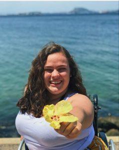 #paratodosverem: Com o mar ao fundo, Ana Laura sorri, segurando uma flor amarela. Foto: arquivo pessoal.